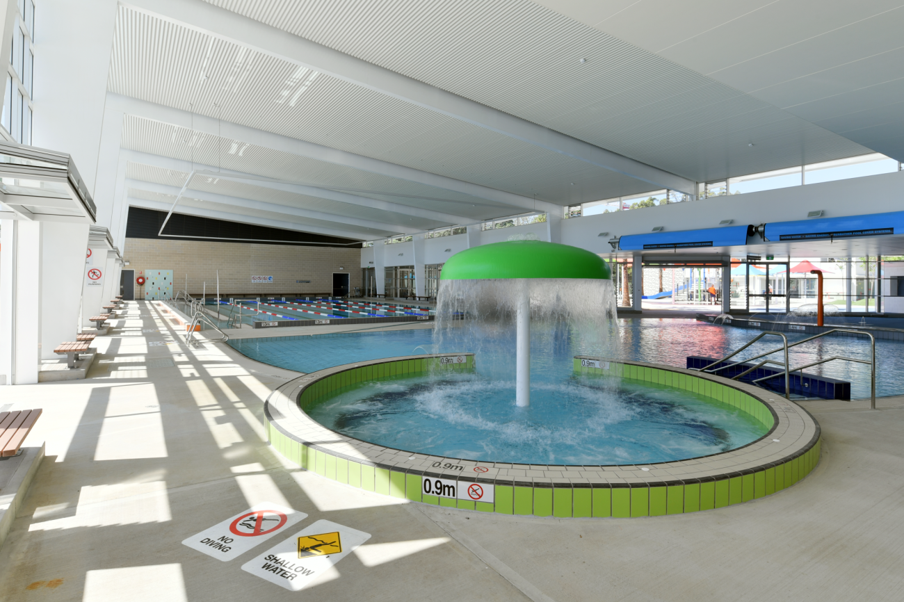 Interior of an aquatic centre featuring a children's mushroom-shaped water feature in the foreground, a large pool in the background, and PERMALITE® roof sheeting in Surfmist® on the roof.